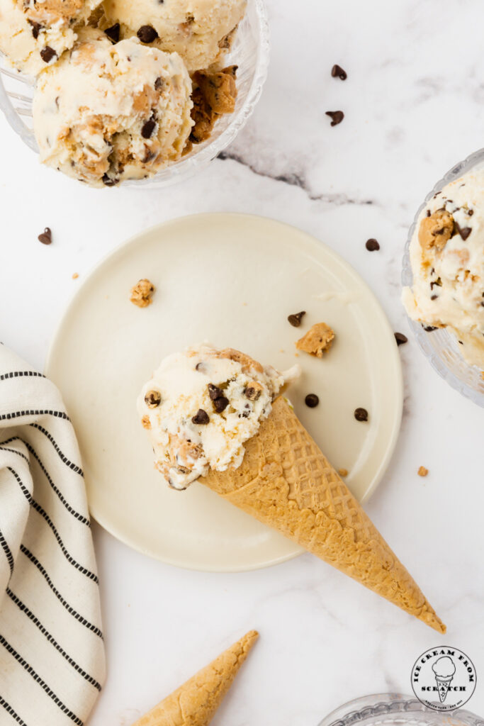 a sugar cone topped with a scoop of cookie dough ice cream, set down on a round white plate.