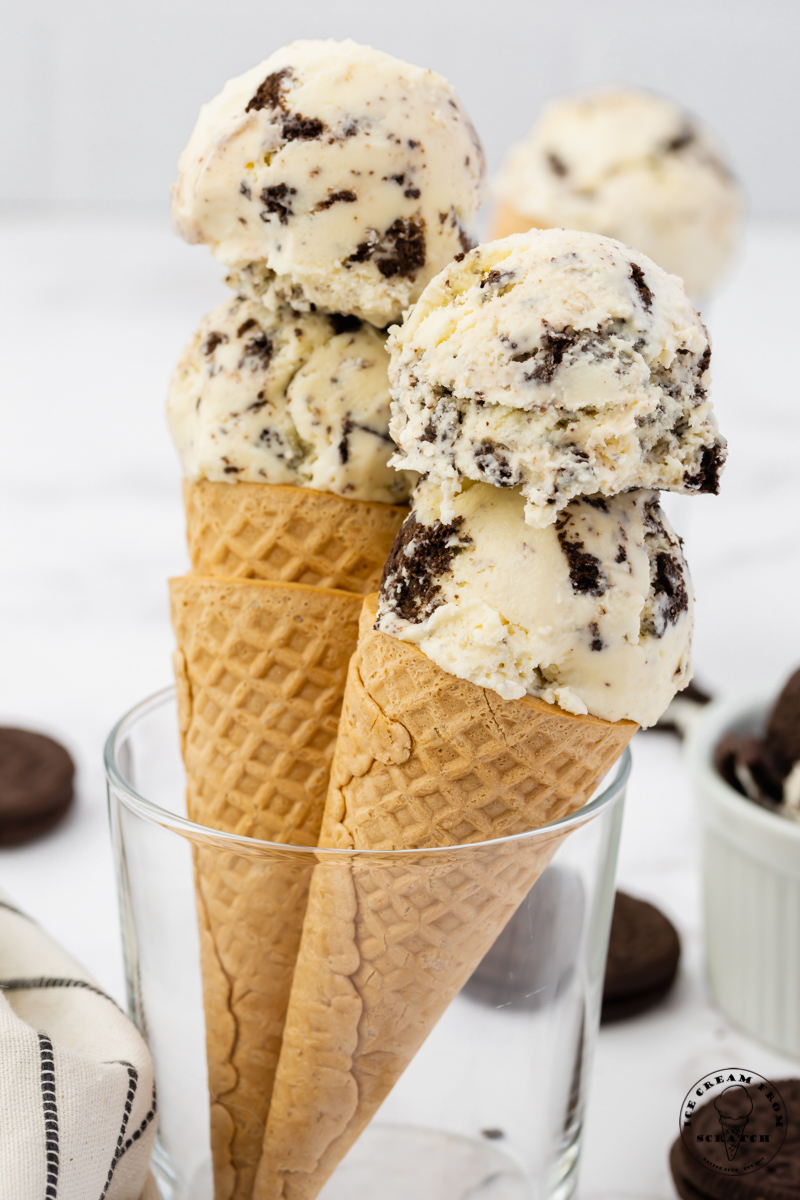 Two sugar cones with double scoops of Oreo cookies and cream ice cream held up in a glass