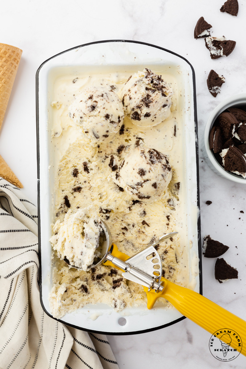 a white enameled rectangular dish filled with homemade Oreo ice cream that is being scooped with a yellow handled ice cream scoop. View from above