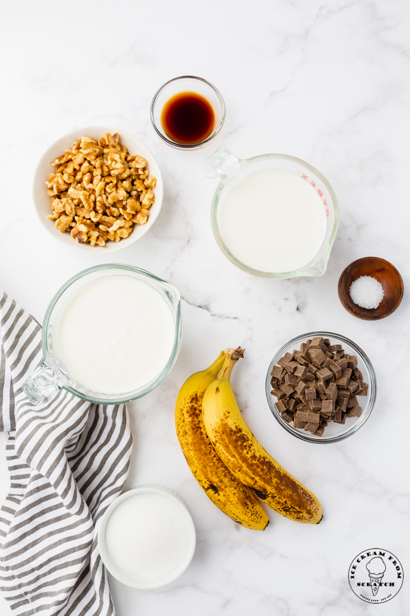 The ingredients for chunky monkey ice cream, including bananas, chocolate, and walnuts, all measured into separate bowls and viewed from above on a marble counter.