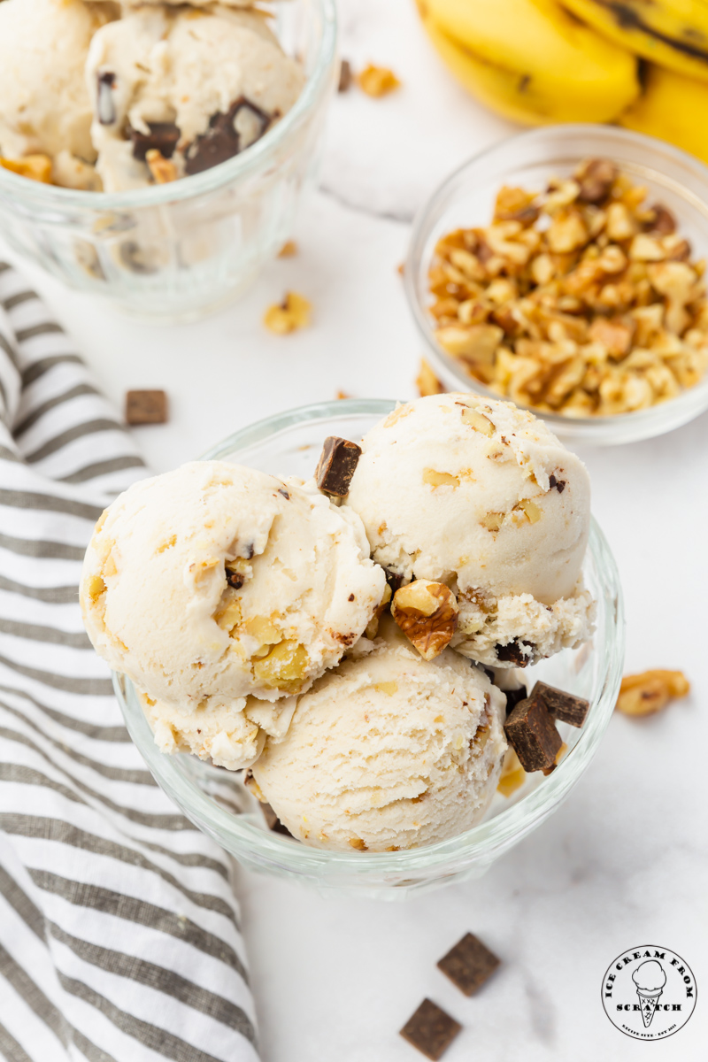 a glass bowl filled with 4 scoops of homemade chunky monkey ice cream topped with chocolate chunks and walnuts. a small bowl of walnuts is in the background next to a bunch of bananas