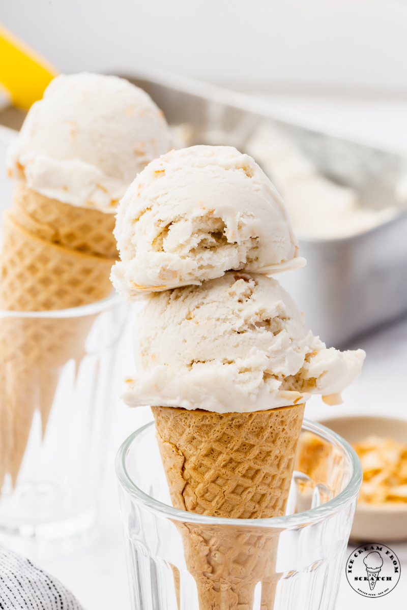 a sugar cone propped up in a glass topped with two scoops of homemade coconut ice cream. another cone sits in the background.