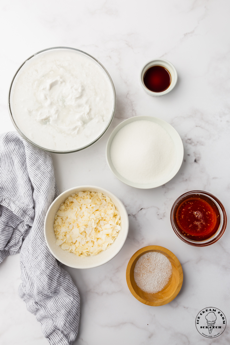The ingredients for coconut ice cream in separate bowls viewed from above on a marble counter, including coconut cream and honey.