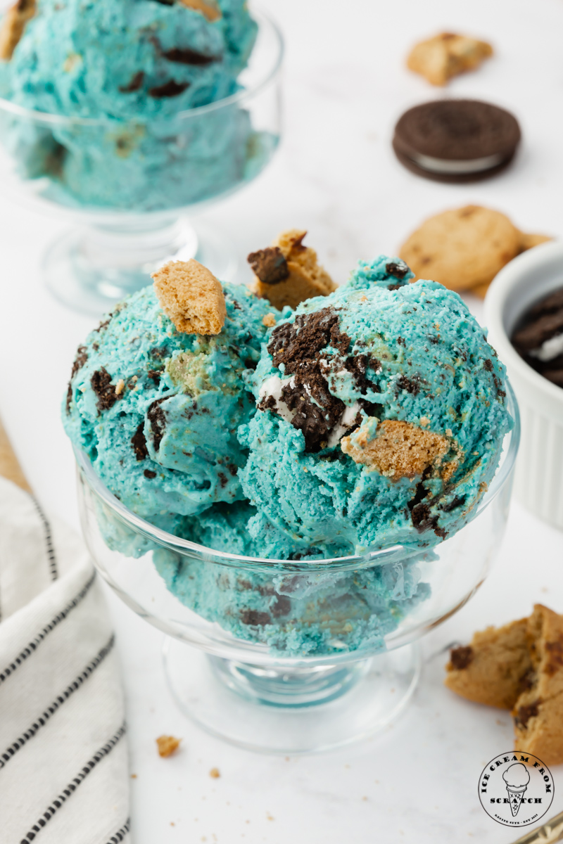 a footed glass ice cream dish filled with three scoops of blue ice cream filled with oreo and chocolate chip pieces