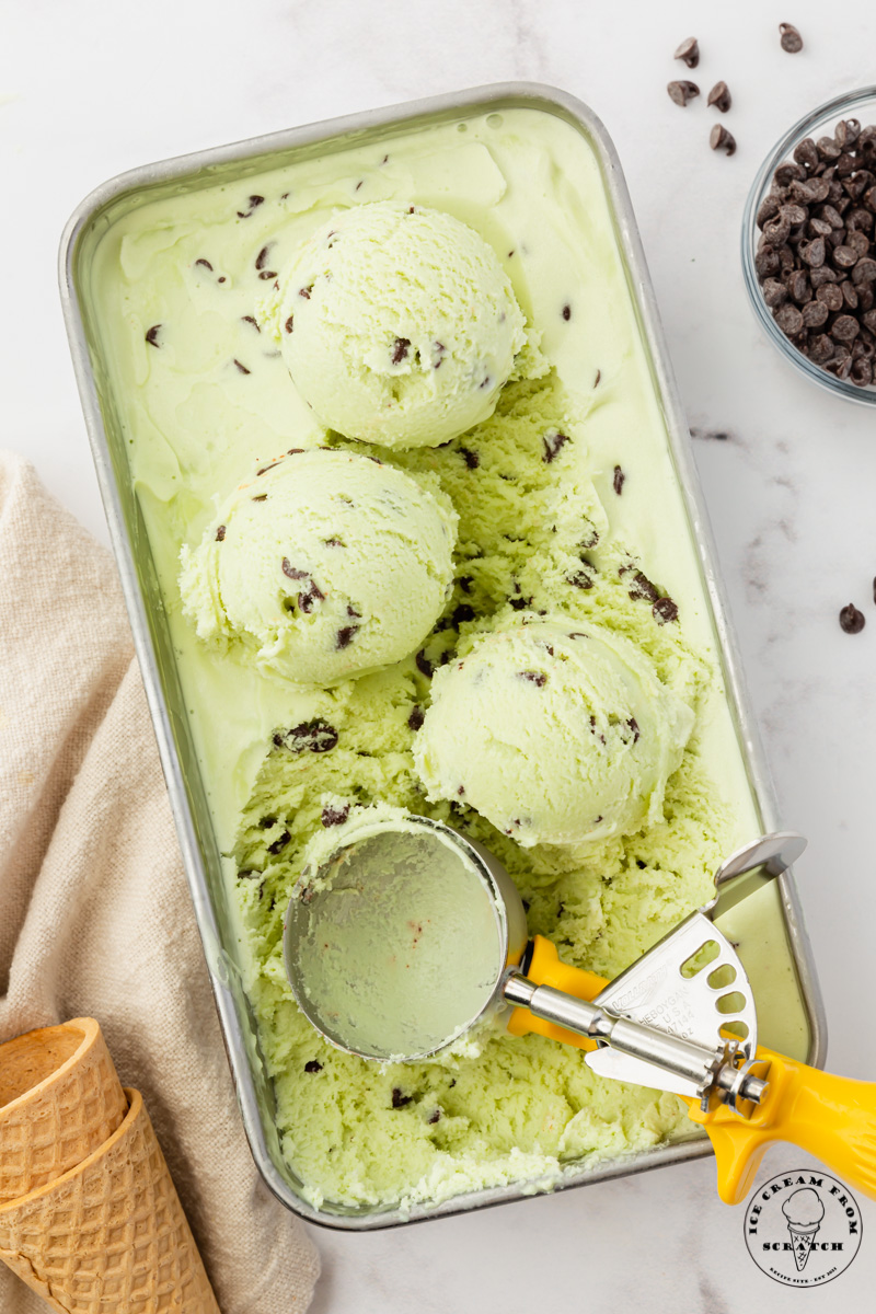 top down view of a rectangular metal pan of homemade mint chocolate chip ice cream. a yellow handled metal ice cream scoop has made three round scoops in the pan.