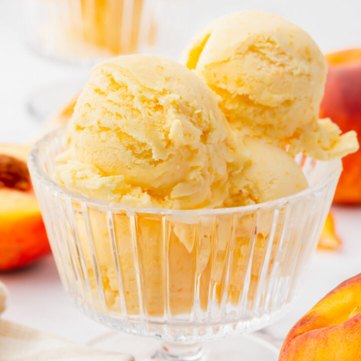 a footed glass ice cream dish filled with 4 scoops of fresh peach ice cream. Peaches are in the background.