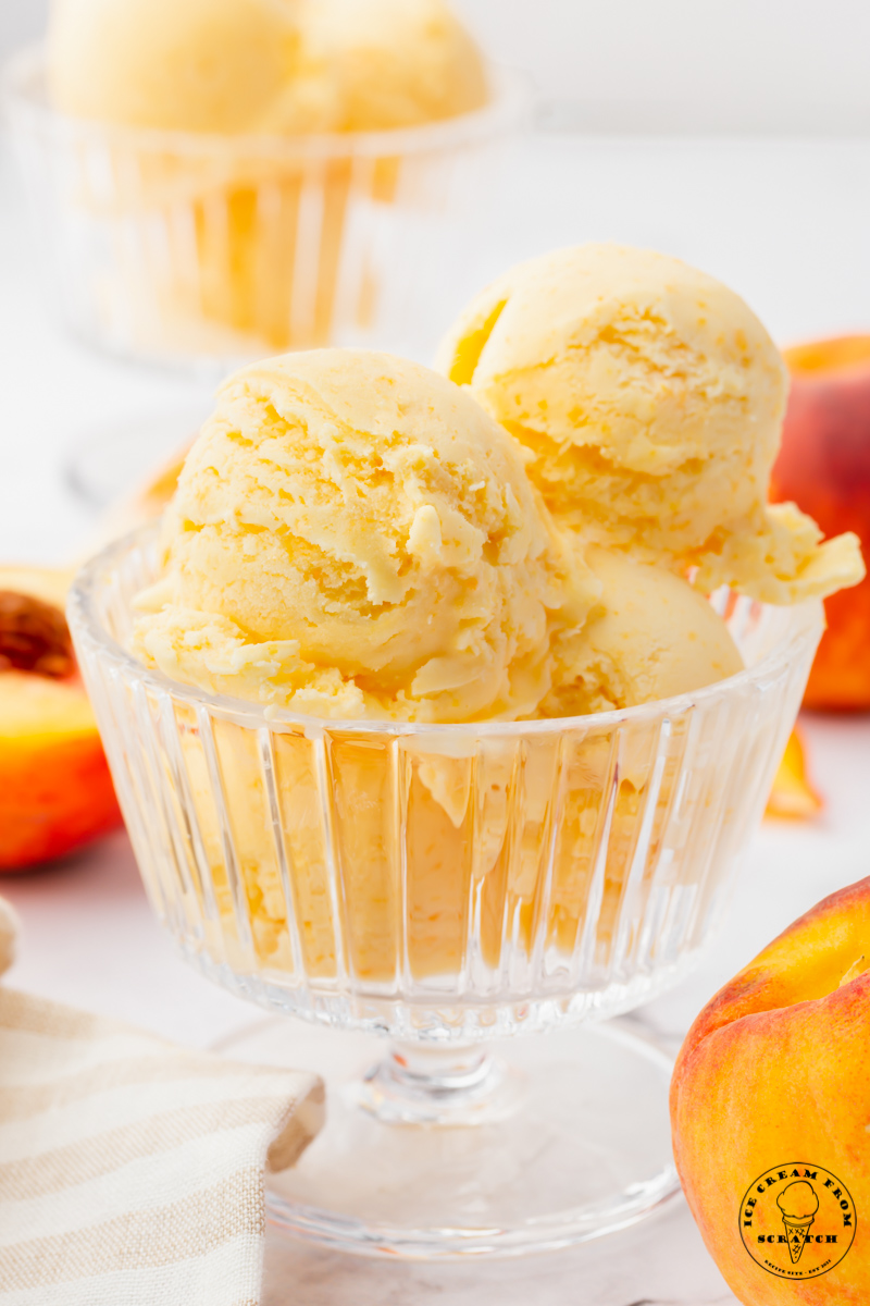 a footed glass ice cream dish filled with 4 scoops of fresh peach ice cream. Peaches are in the background.