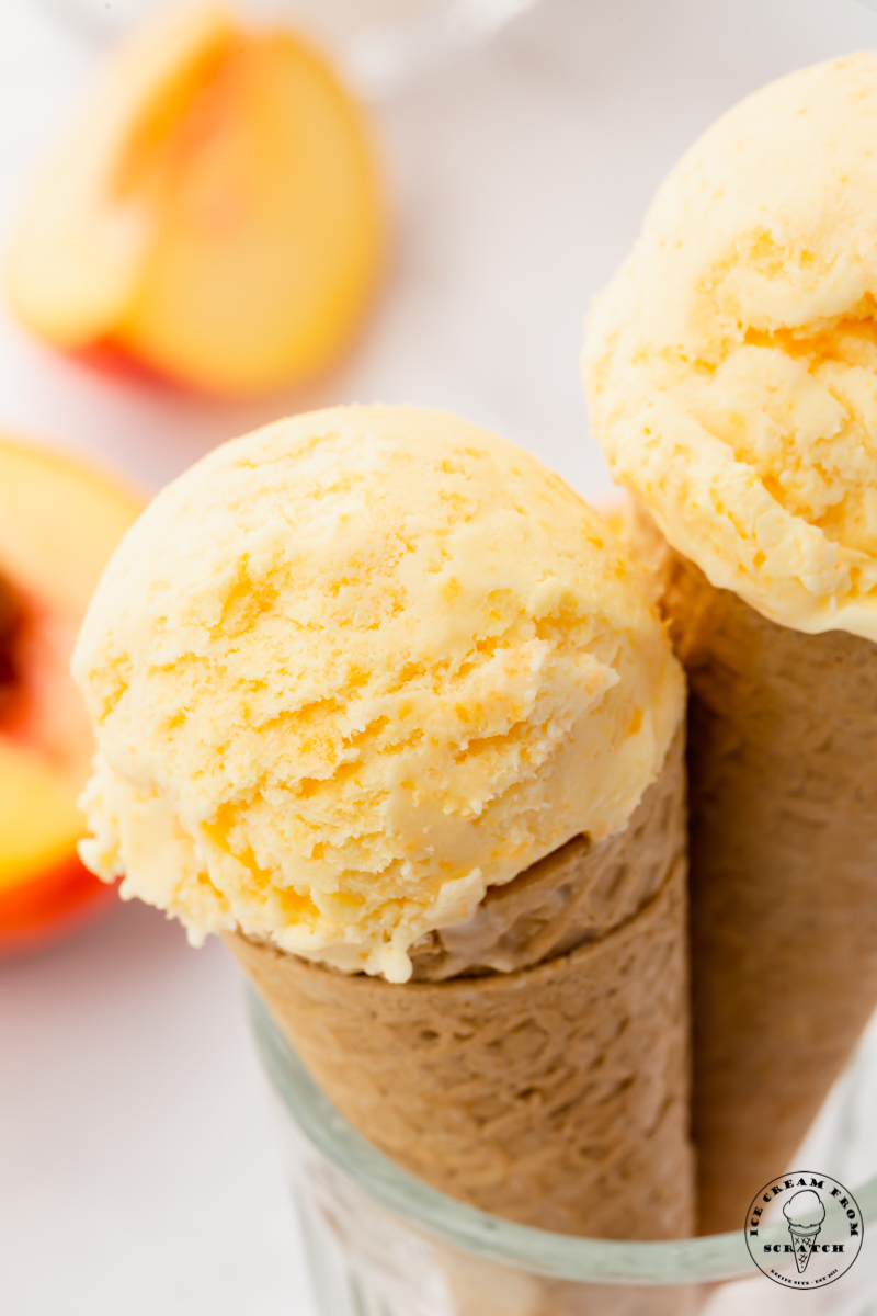 two sugar cones topped with a scoop of fresh peach ice cream, propped up in a glass.