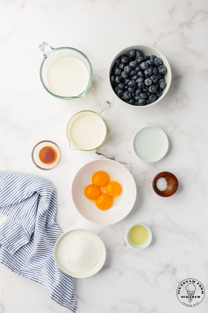 The ingredients for fresh, homemade blueberry ice cream measured into bowls on a marble counter, viewed from above