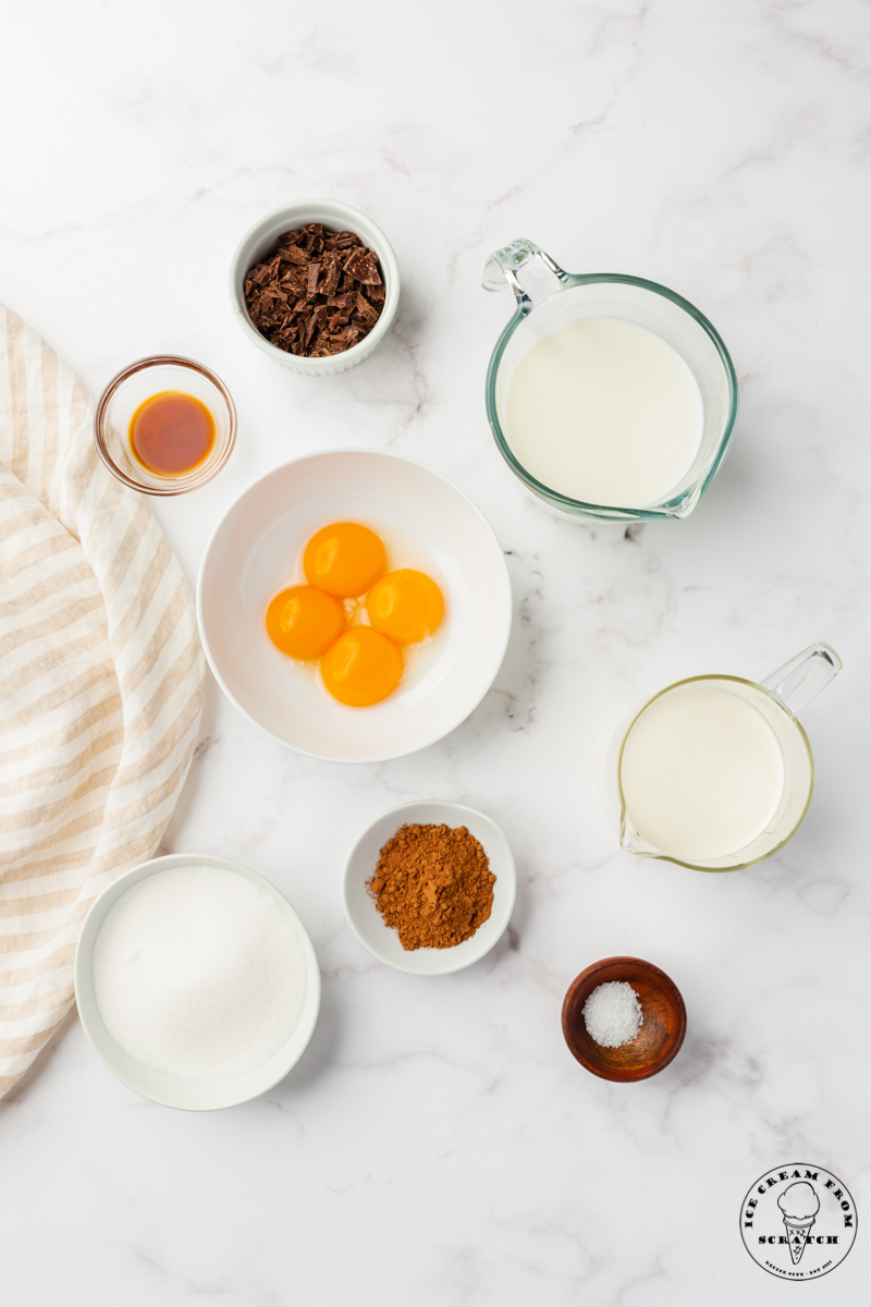 The ingredients for homemade chocolate ice cream, measured out into separate bowls on a marble counter, including egg yolks, cream, cocoa, chopped chocolate, and sugar.