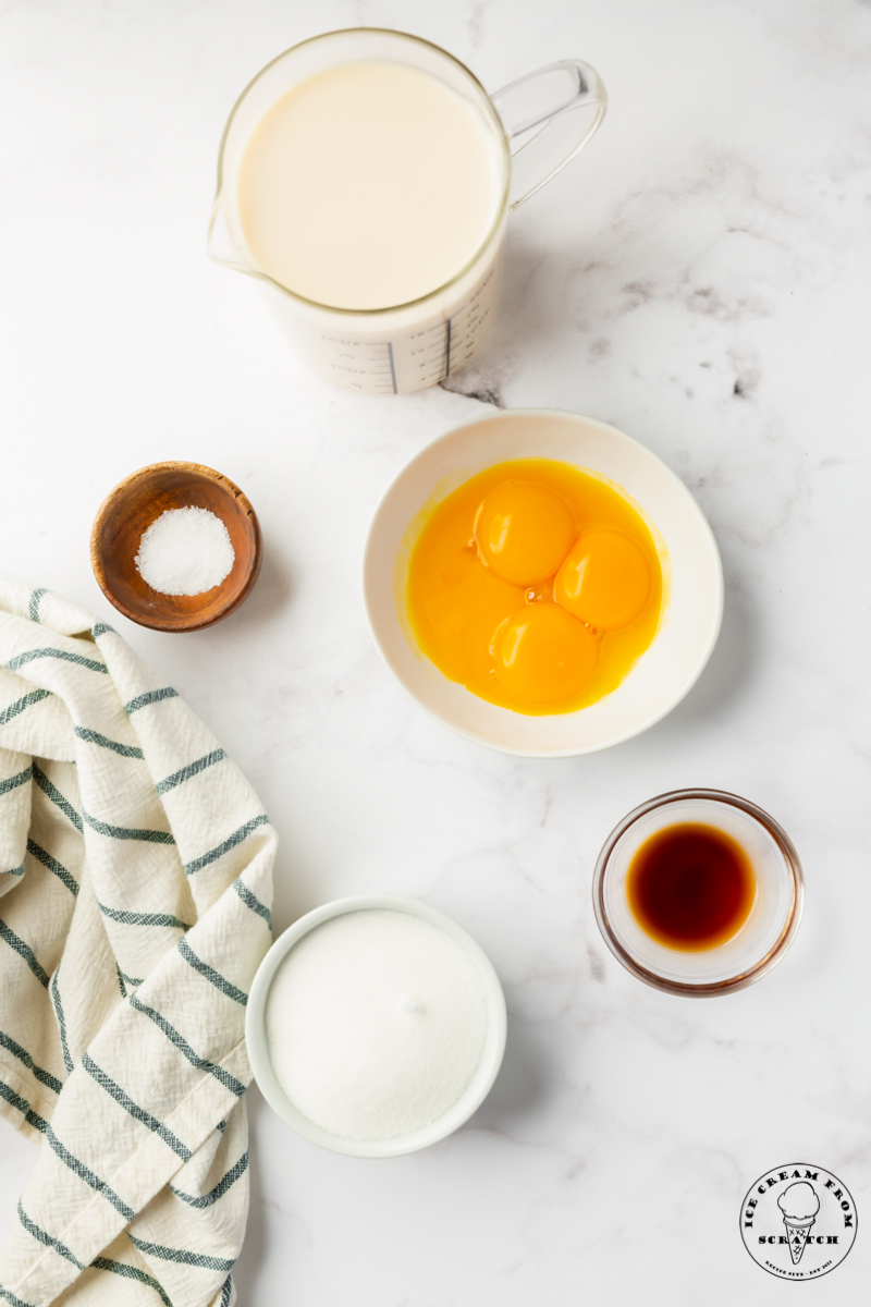 The 5 ingredients needed to make homemade oat milk ice cream, measured into separate bowls on a marble countertop next to a linen striped towel.
