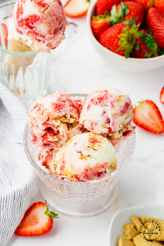 a glass dish filled with three scoops of strawberry cheesecake ice cream packed with berries and graham cracker pieces. In the background is a bowl of strawberries and a cone of the same ice cream.