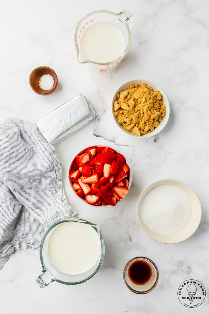 The ingredients for strawberry cheesecake ice cream recipe, including cream cheese, graham cracker pieces, and fresh strawberries. Each ingredient is measured into a separate bowl on a marble counter, viewed from above.