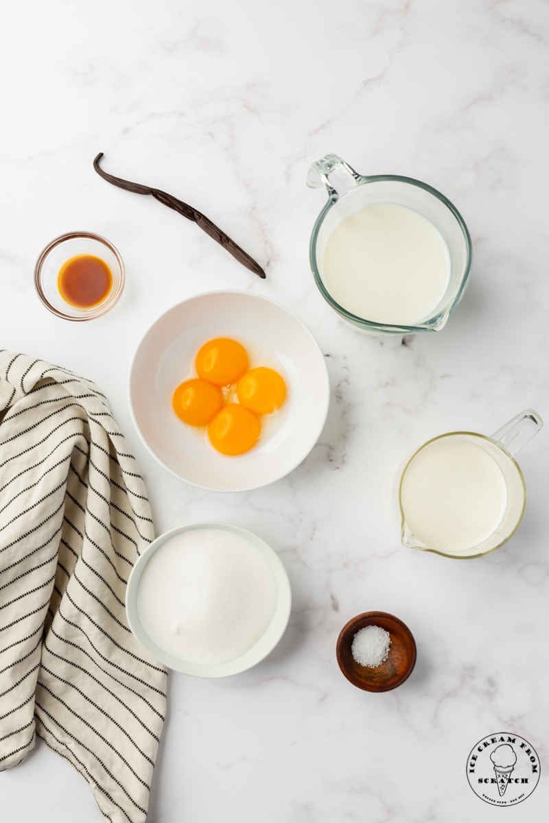The simple, natural ingredients for making homemade vanilla ice cream, including a whole vanilla bean, egg yolks, and other ingredients, in separate bowls, viewed from above.