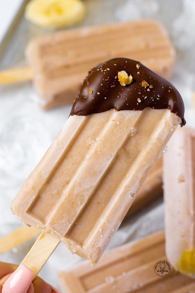 a light brown banana popsicle with just the top dipped in chocolate, sprinkled with chopped walnuts
