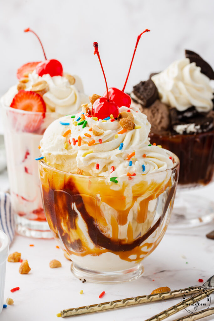 A glass bowl filled with a chocolate caramel ice cream sundae, topped with whipped cream, sprinkles, and two maraschino cherries. More ice cream sundaes of different varieties are in the background.