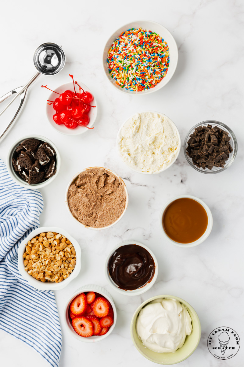 The ingredients needed to create an ice cream sundae bar, all in small bowls on a marble countertop, viewed from above.