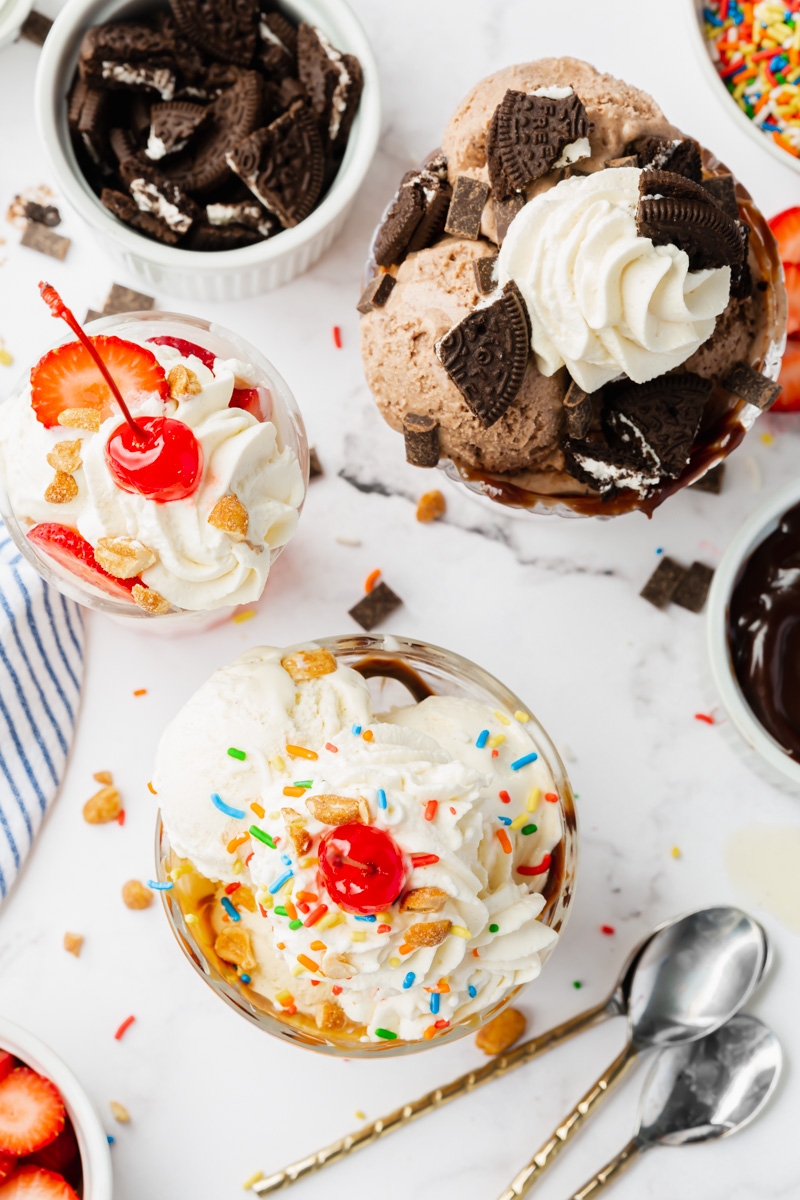 Three different ice cream sundaes, one classic with whipped cream and cherries, one strawberry, and one chocolate with crushed oreos.