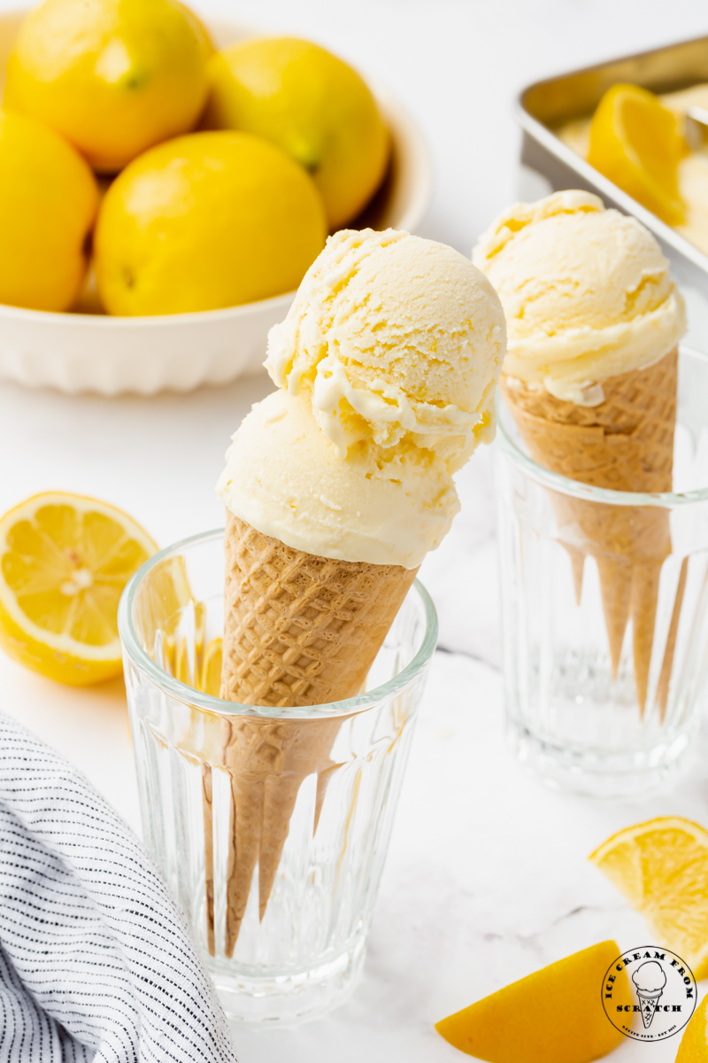 Two sugar cones topped with light yellow lemon ice cream, being held up in glass drinking glasses. A bowl of lemons is in the background.