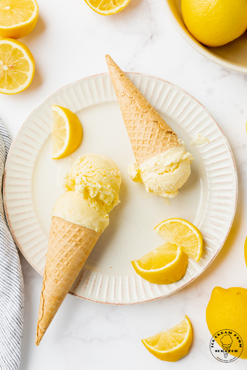 a white fluted plate holding two sugar cones of homemade lemon ice cream, garnished with lemon wedges. Viewed from above.