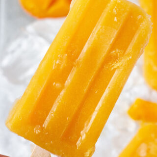 an orange colored mango popsicle on a wooden stick, held by a feminine hand, up close.