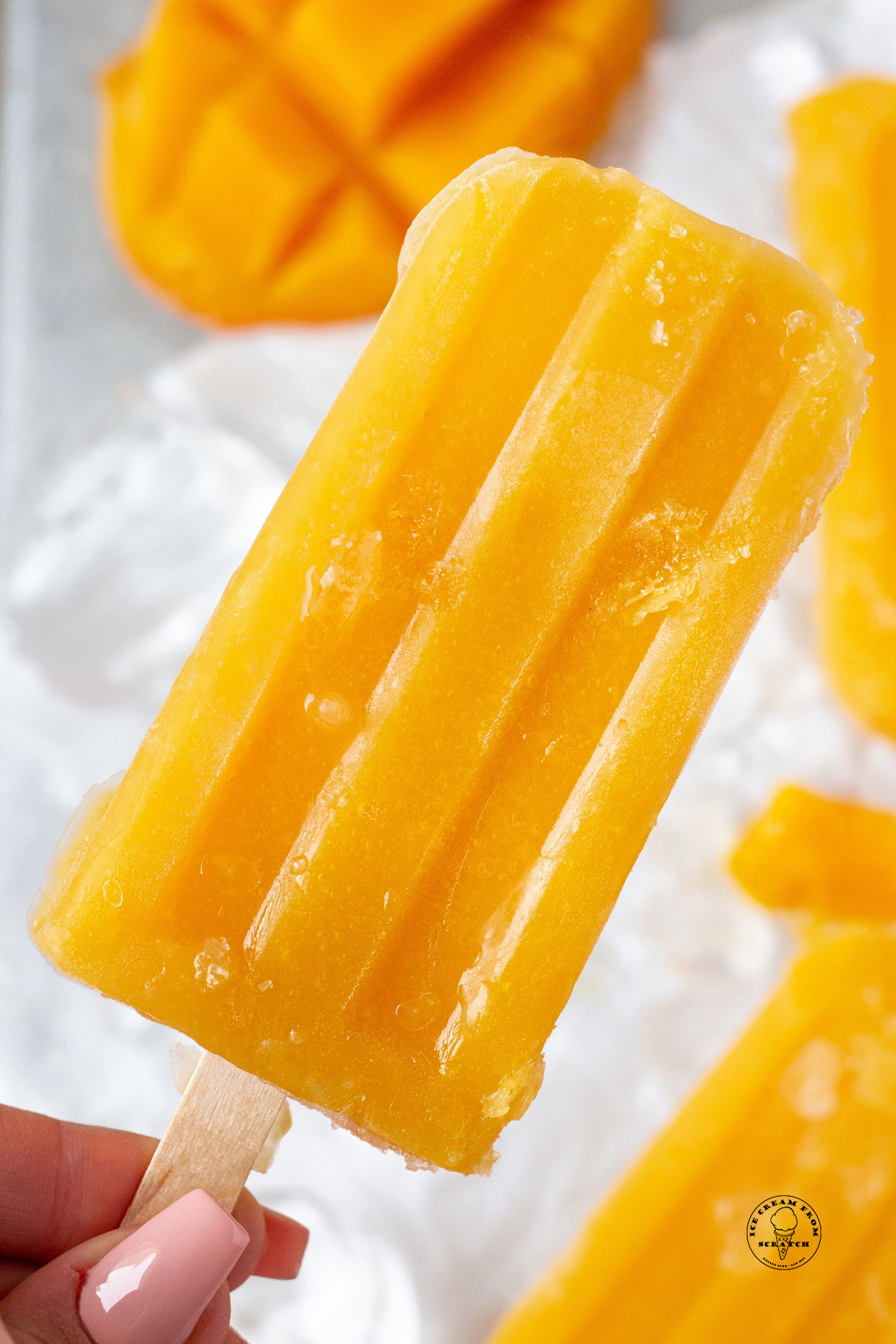 an orange colored mango popsicle on a wooden stick, held by a feminine hand, up close.