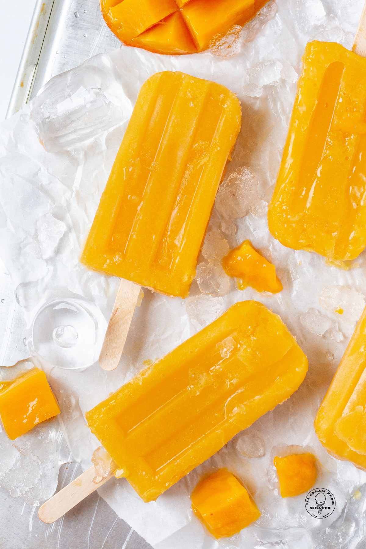 Four mango popsicles on a tray of ice. Small chunks of mango are next to them.