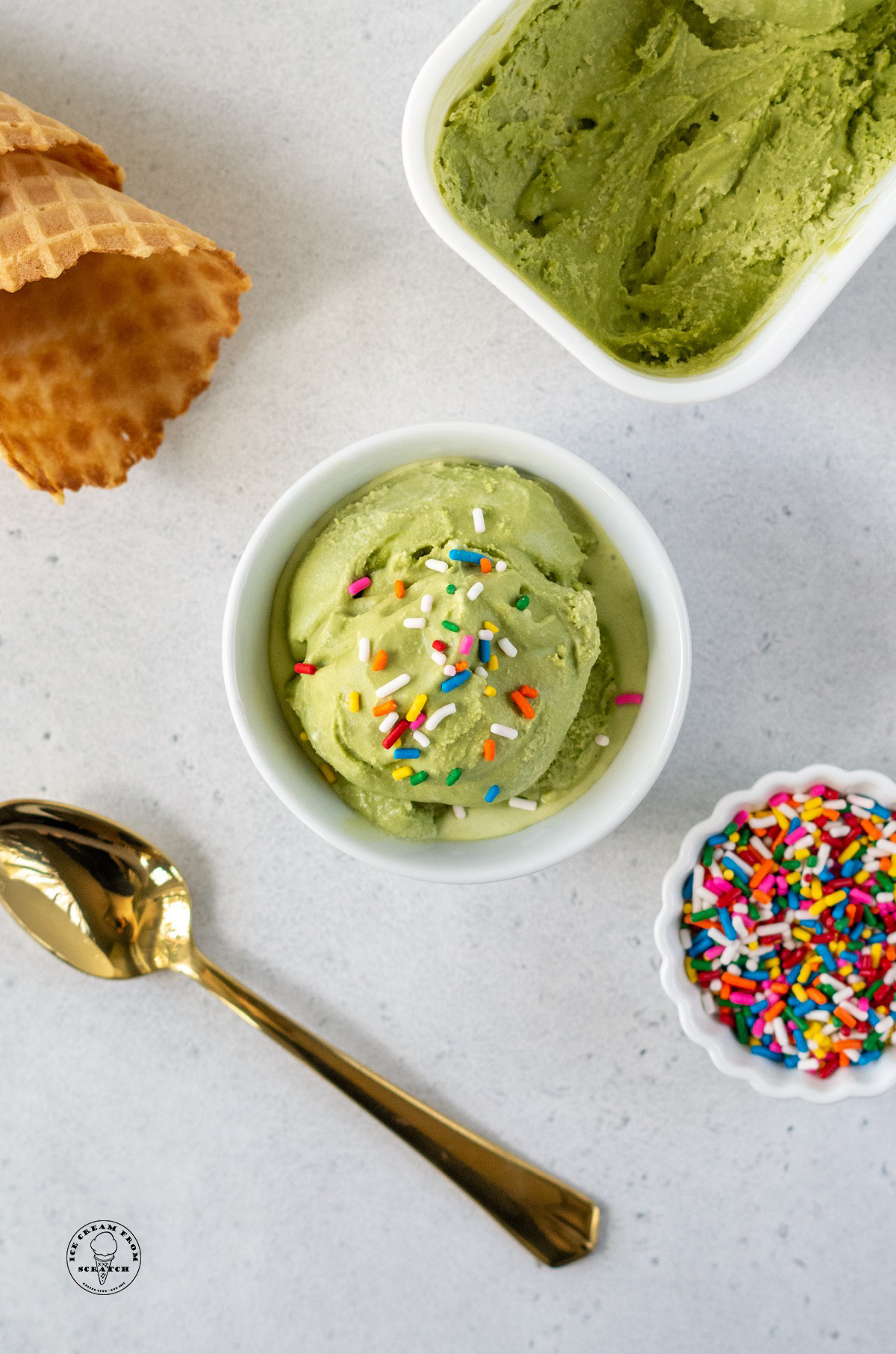 a small white bowl with a scoop of homemade matcha ice cream topped with rainbow sprinkles, viewed from above. Around the ice cream are waffle cones, a gold spoon, and a bowl of sprinkles