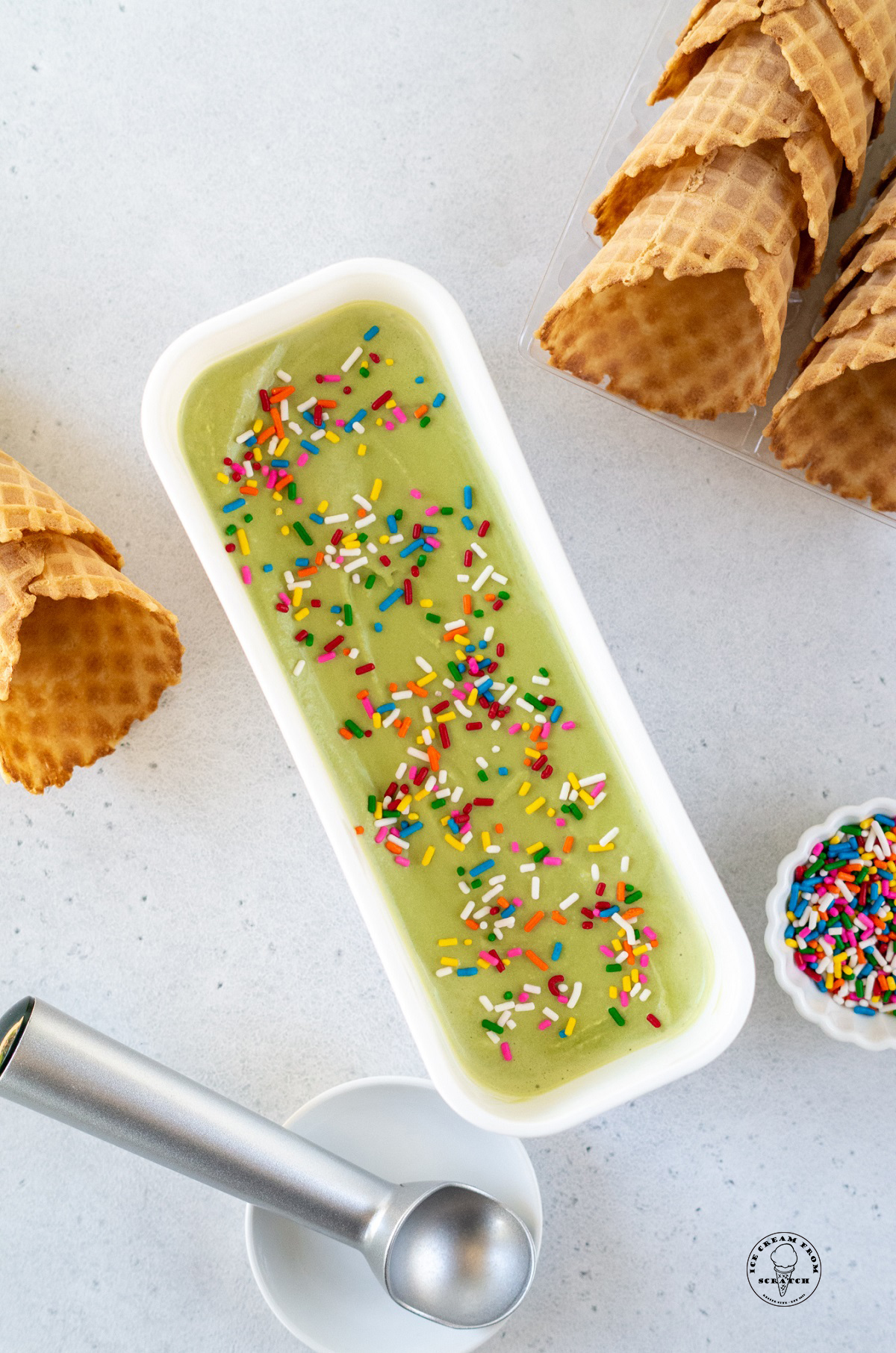 a narrow, long white, rectangular container filled with green matcha ice cream next to rows of waffle cones. Viewed from above