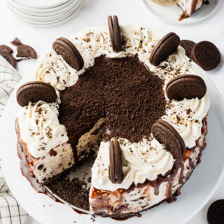 a large homemade ice cream cake topped with whipped cream and whole oreo cookies, stood up around the edge. The cake is on a plate. Behind it is a small plate with a slice of oreo ice crema cake, and a stack of small plates and two gold forks.
