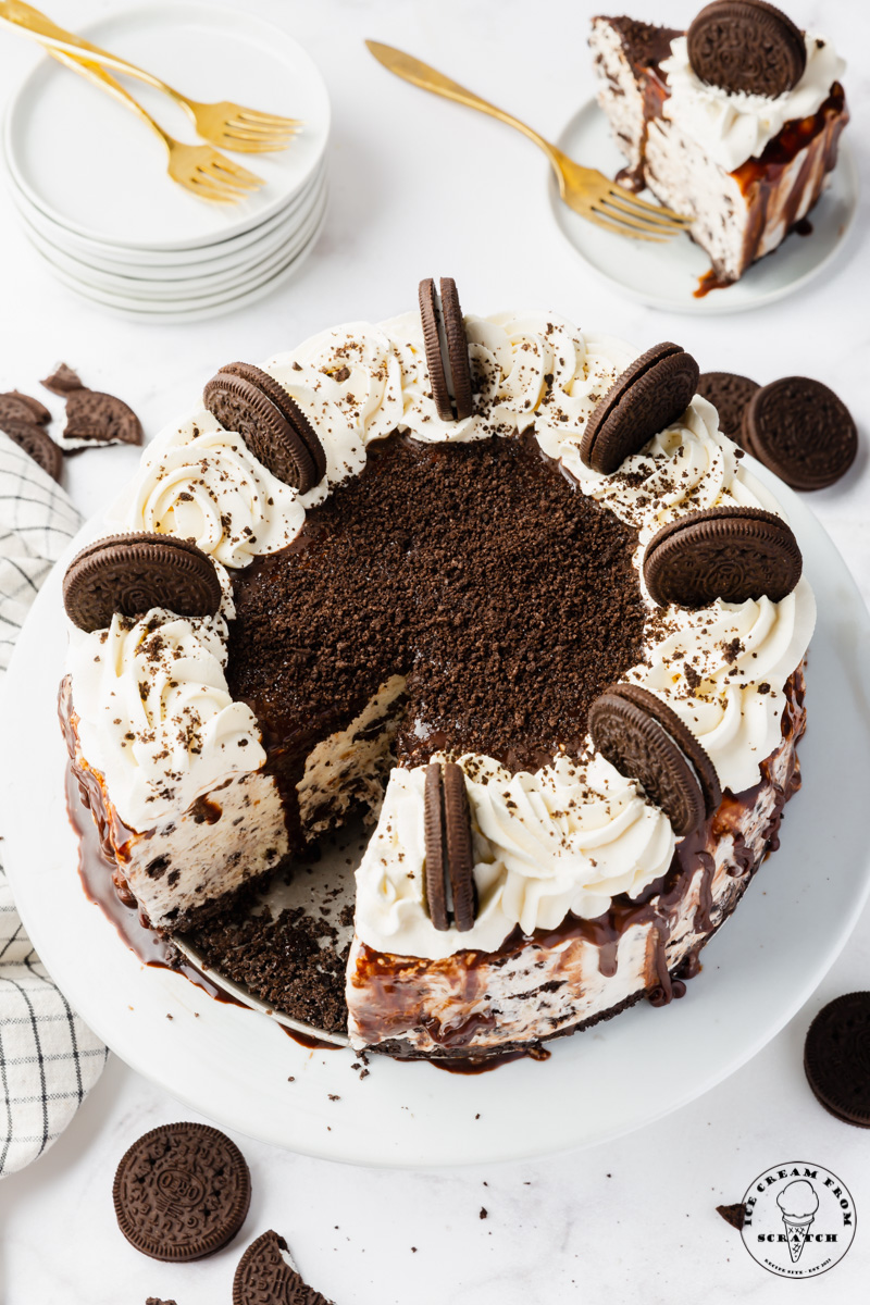 a large homemade ice cream cake topped with whipped cream and whole oreo cookies, stood up around the edge. The cake is on a plate. Behind it is a small plate with a slice of oreo ice crema cake, and a stack of small plates and two gold forks.