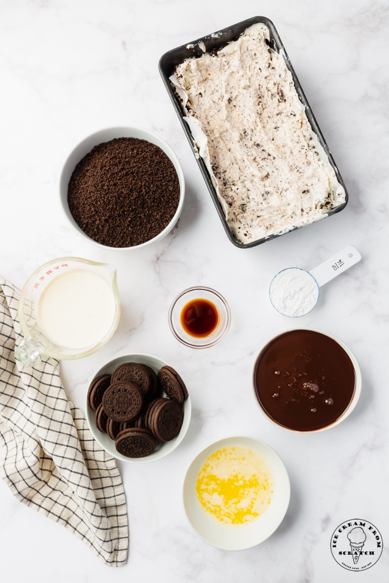 The ingredients needed for making an oreo ice cream cake, including homemade oreo ice cream, chocolate sauce, and crushed oreos, all in separate bowls on a marble countertop.