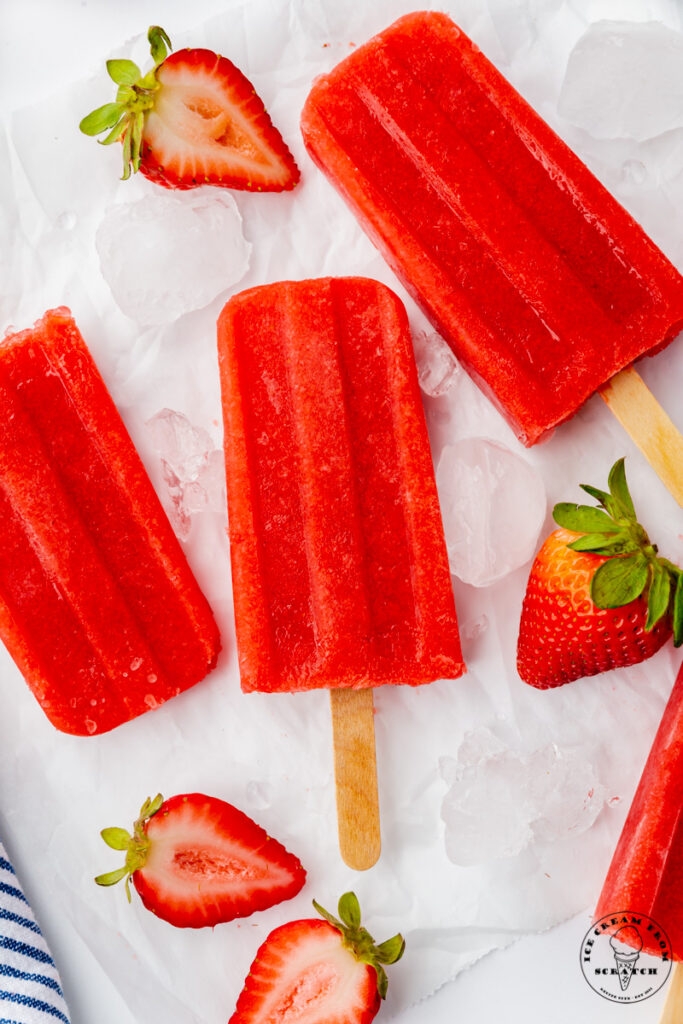 three strawberry popsicles on top of ice cubes with halved strawberries, viewed from above.