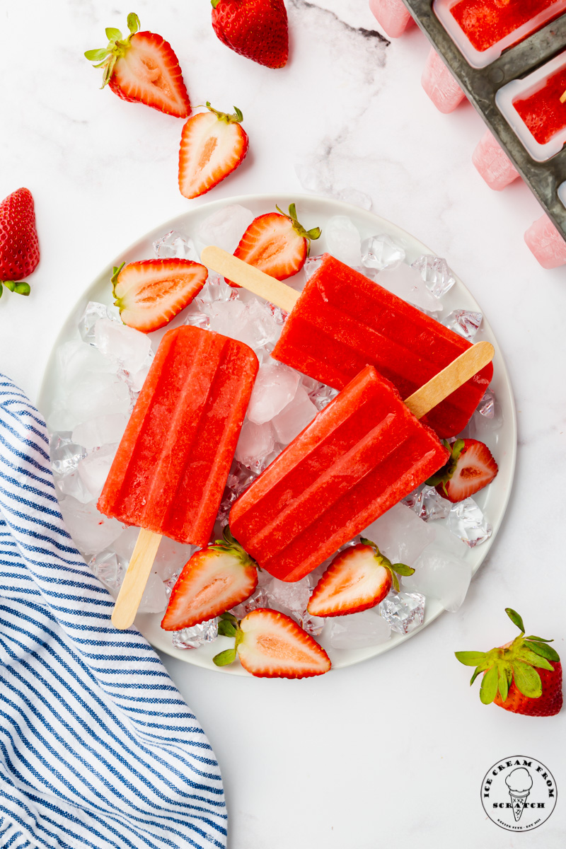 A round plate of ice topped with three homemade strawberry popsicles and halved berries.