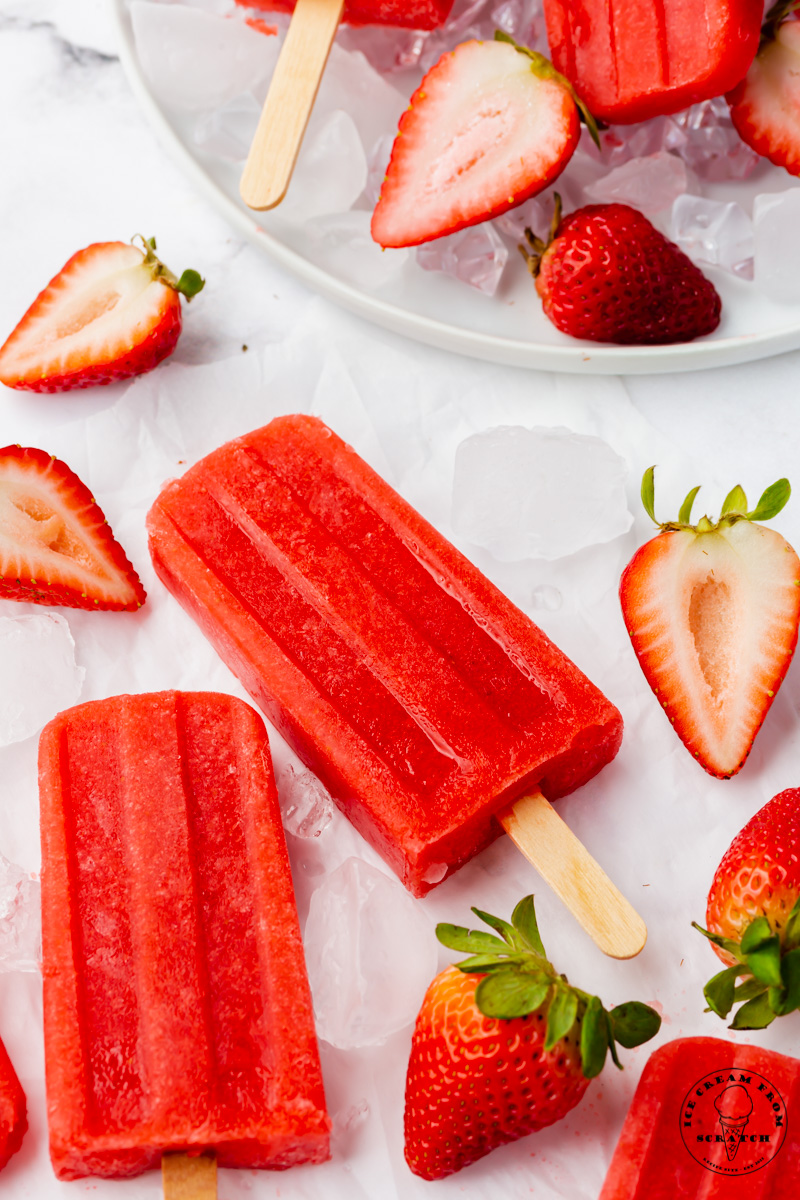 Two strawberry popsicles and fresh berries laid on a bed of ice cubes.