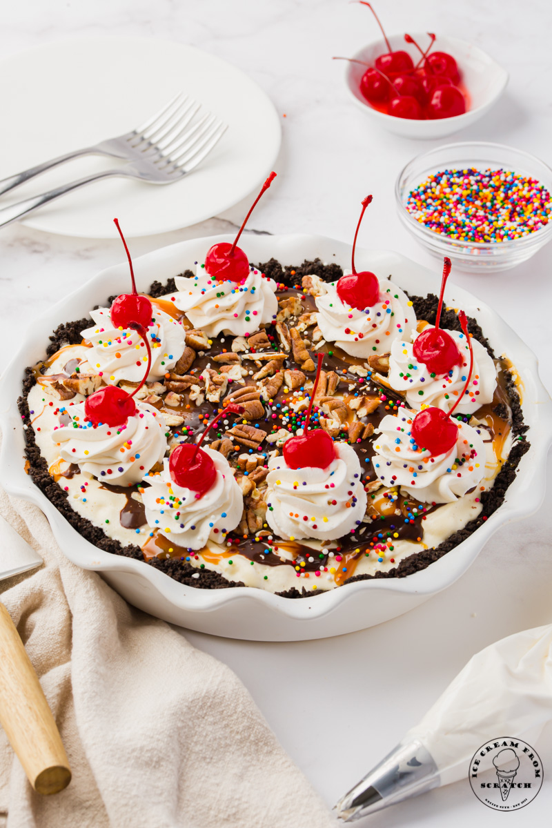 a homemade ice cream pie topped with piles of whipped cream, cherries, sprinkles, and pecans.