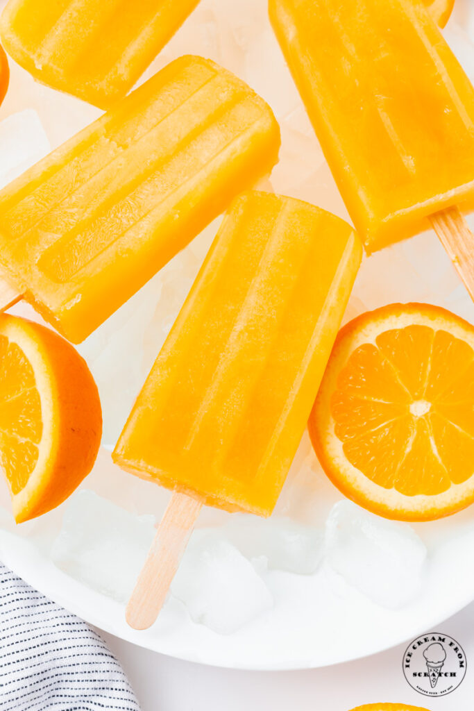 a plate of ice, holding rectangular orange popsicles with wooden sticks and sliced oranges.