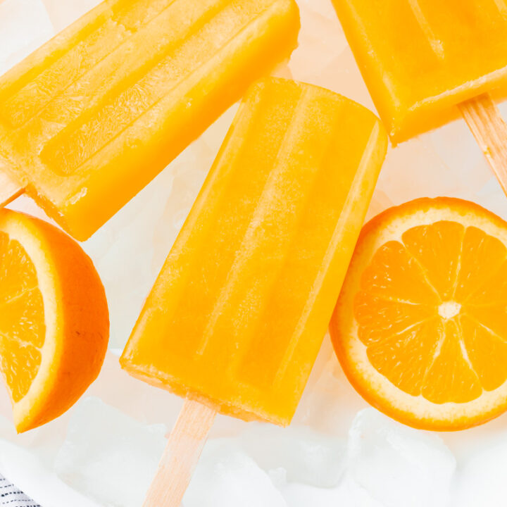 a plate of ice, holding rectangular orange popsicles with wooden sticks and sliced oranges.