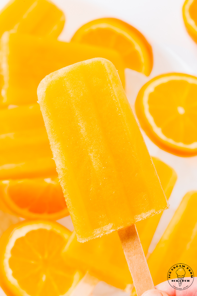 a rectangle orange popsicle on a wooden stick, held up above a pile of orange slices.