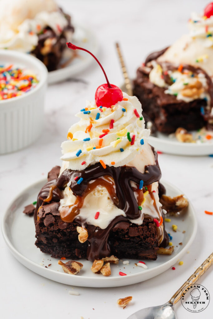 a brownie sundae on a small white plate, topped with ice cream, hot fudge, caramel whipped cream, chopped walnuts, rainbow sprinkles, and a cherry on top.