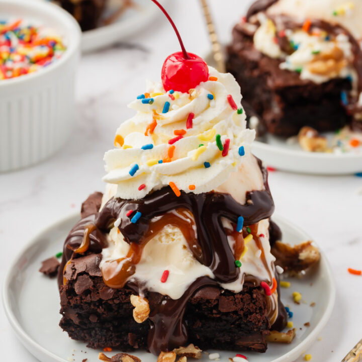 a brownie sundae on a small white plate, topped with ice cream, hot fudge, caramel whipped cream, chopped walnuts, rainbow sprinkles, and a cherry on top.