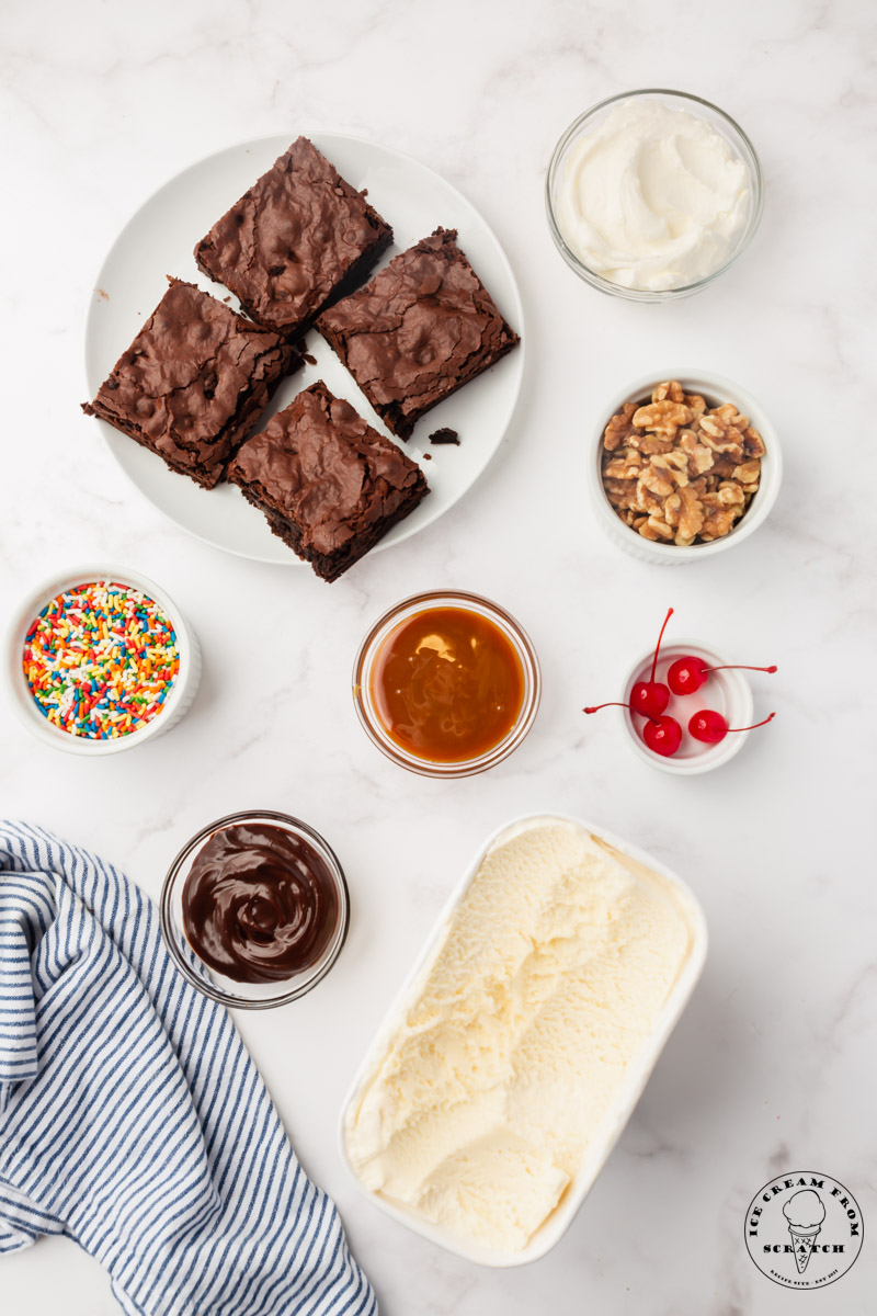 The ingredients to make homemade brownie sundaes, all in small bowls, arranged on a marble countertop, and viewed from above. There is a plate of four brownies, a box of vanilla ice cream, and small bowls of sprinkles, nuts, whipped cream, maraschino cherries with stems, fudge sauce, and caramel topping.