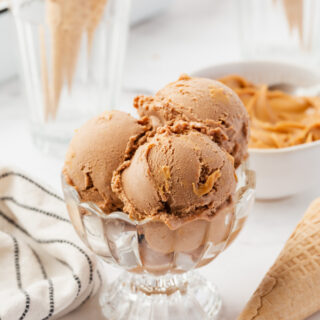 a footed glass dessert dish with three scoops of peanut butter swirled chocolate ice cream. Cones and a bowl of peanut butter are in the background.