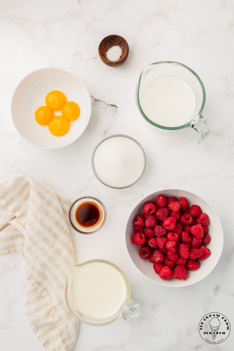 The ingredients needed to make french style homemade raspberry ice cream, all in small bowls on a marble countertop, viewed from above
