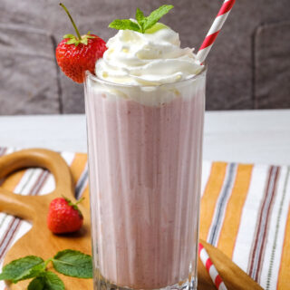 a strawberry milkshake topped with whipped cream, mint leaves, and a fresh berry. A red and white striped straw is inserted, and the shake sits on top of a small wooden board.
