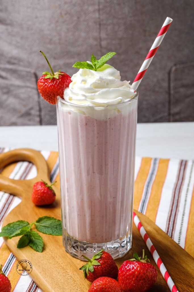 a strawberry milkshake topped with whipped cream, mint leaves, and a fresh berry. A red and white striped straw is inserted, and the shake sits on top of a small wooden board.