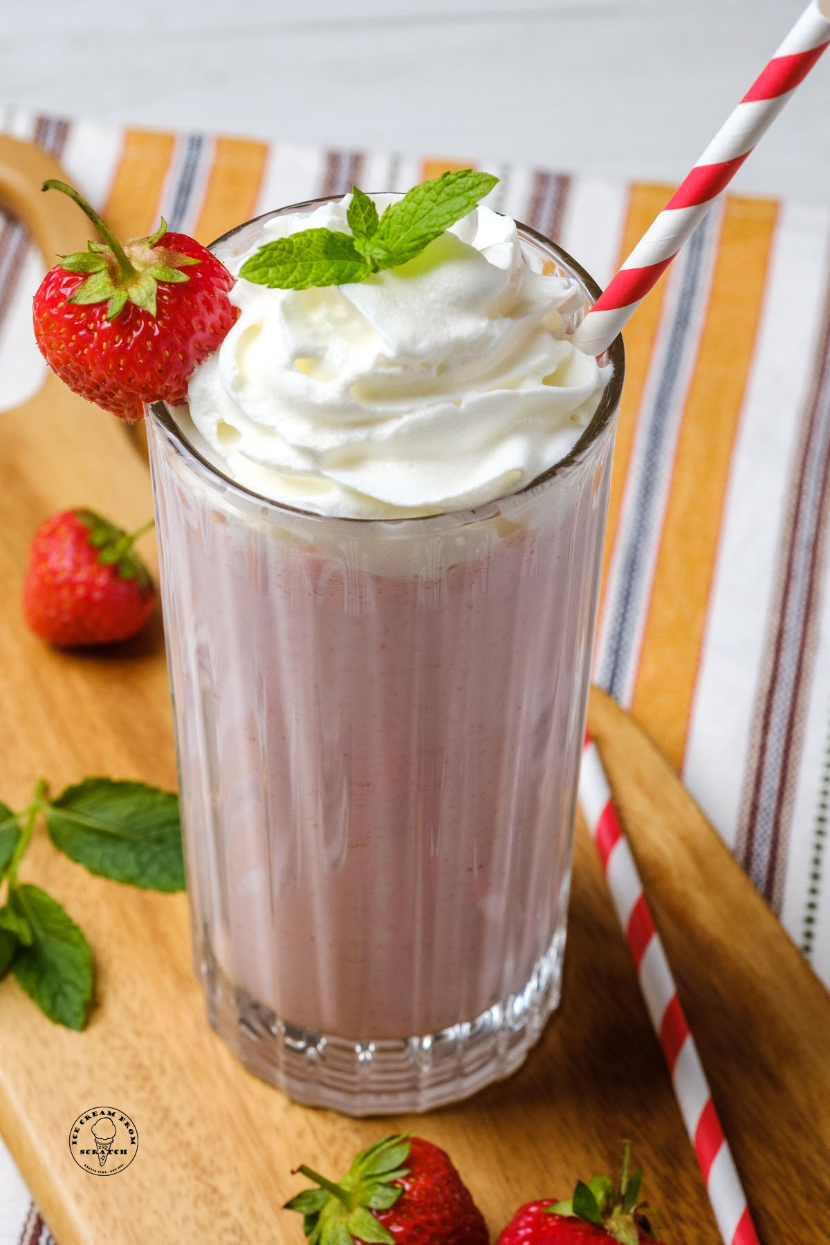 a ridged clear glass filled with strawberry milkshake, garnished with whipped ream, mint, strawberries, and a red and white straw.