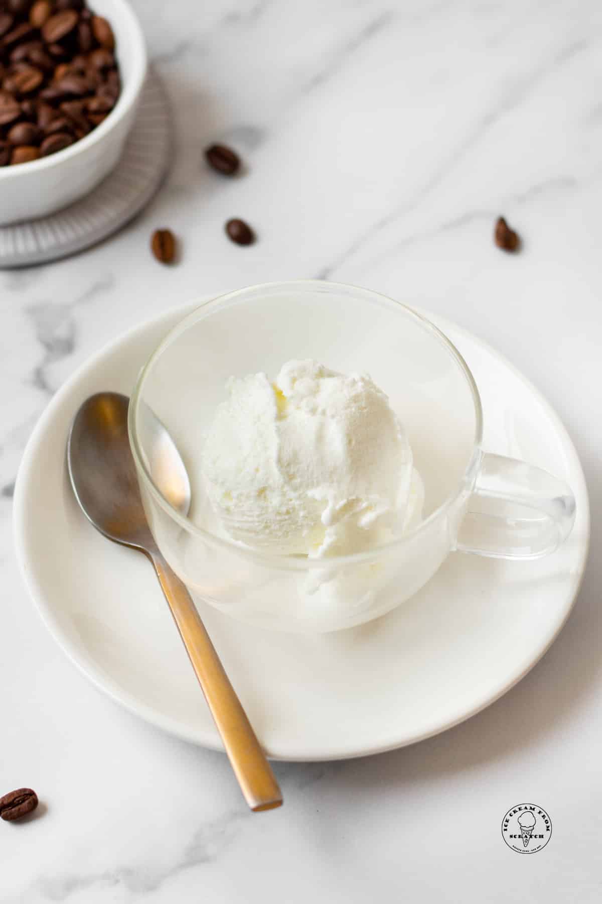 a clear coffee mug filled with a scoop of vanilla ice cream, on a saucer with a teaspoon.