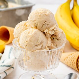 scoops of homemade banana foster ice cream in a footed glass dish, garnished with a slice of banana. Ice Cream cones and yellow bananas are in the background.