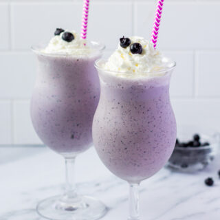 Two blueberry milkshakes in hurricane glasses with purple and white striped straws